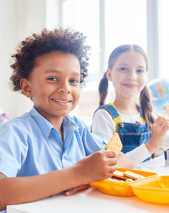food-allergy-training-for-childcare Two kids smiling while eating lunch in a cafeteria.